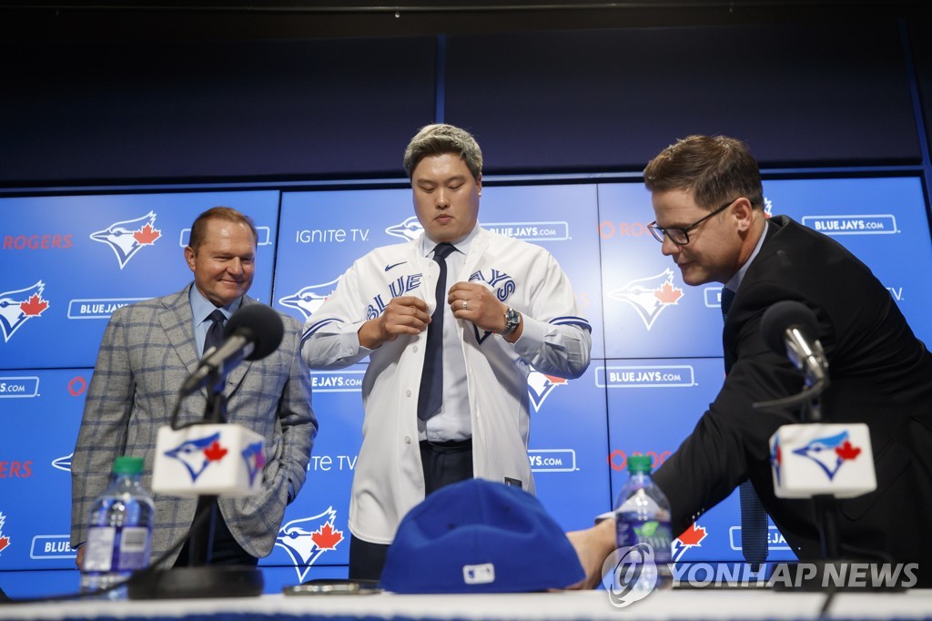 In this Canadian Press photo via the Associated Press from Dec. 27, 2019, Ryu Hyun-jin (C) puts on his new jersey for the Toronto Blue Jays during his introductory press conference at Rogers Centre in Toronto. Ryu is flanked by his agent Scott Boras (L) and Blue Jays' general manager Ross Atkins. (Yonhap)