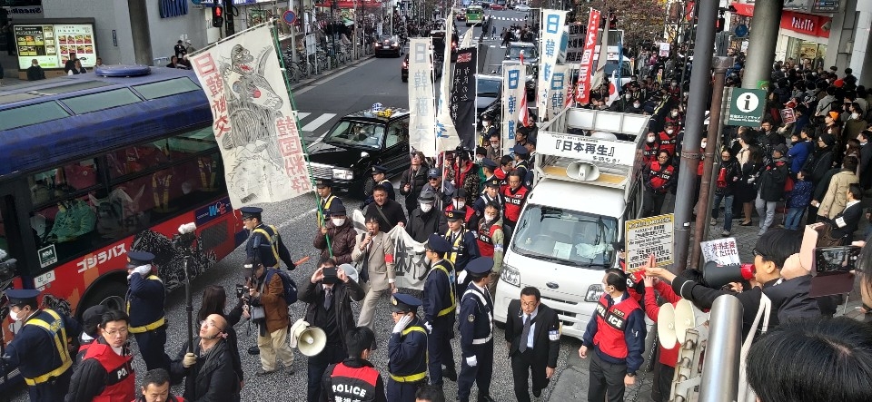 일본 극우단체 회원들이 1일 오후 도쿄 신주쿠역 주변에서 한일 국교 단절을 주장하며 거리행진을 하고 있다. 흰색 선전차량 뒤로 따르는 약 20명이 우익 집회 참가자이고, 나머지 대부분은 행인과 섞인 '맞불집회' 참가자들이다. 붉은색 조끼 착용자와 제복을 입은 사람은 경찰관.